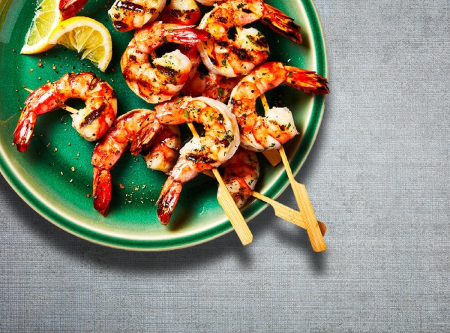 Round green plate with three cooked shrimp skewers laid on top, and cut limes for garnish